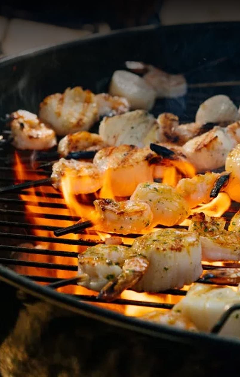 Grilled shrimp and scallop skewers cooking over open flames on a charcoal grill.